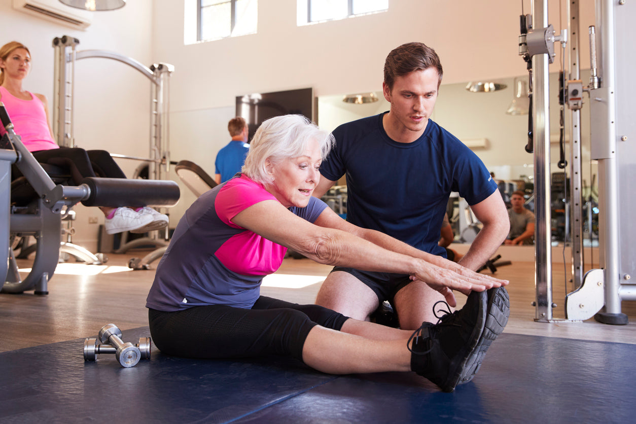 A gym instructor supporting an older woman stretching to touch her toes