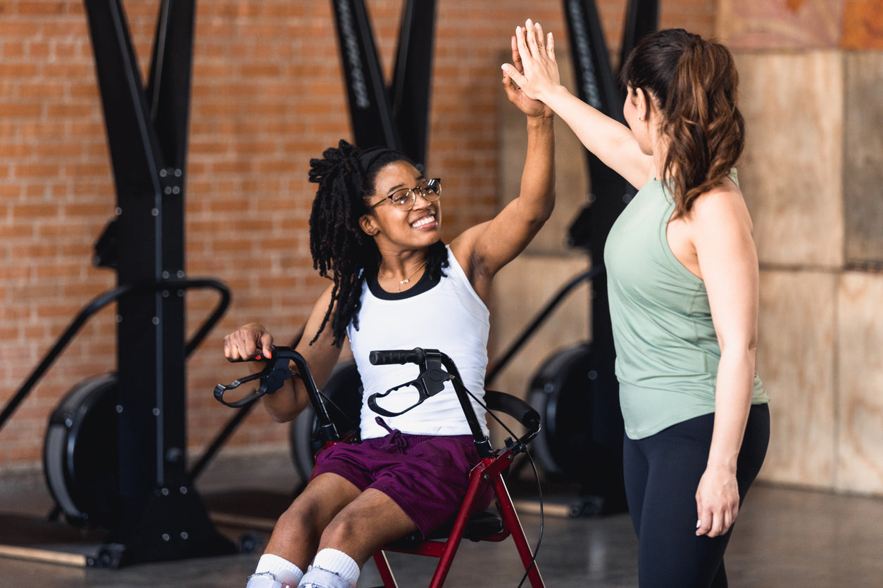 A gym goer in an adapted wheelchair or walker giving a high-five to their personal trainer