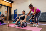 A group of pregnant mothers-to-be taking part in a yoga class in a community centre. They are sitting down on yoga mats with their legs crossed while the instructor of the class teaches them breathing techniques to help with birthing and pregnancy. The instructor is focused on one woman and talking her through the position.