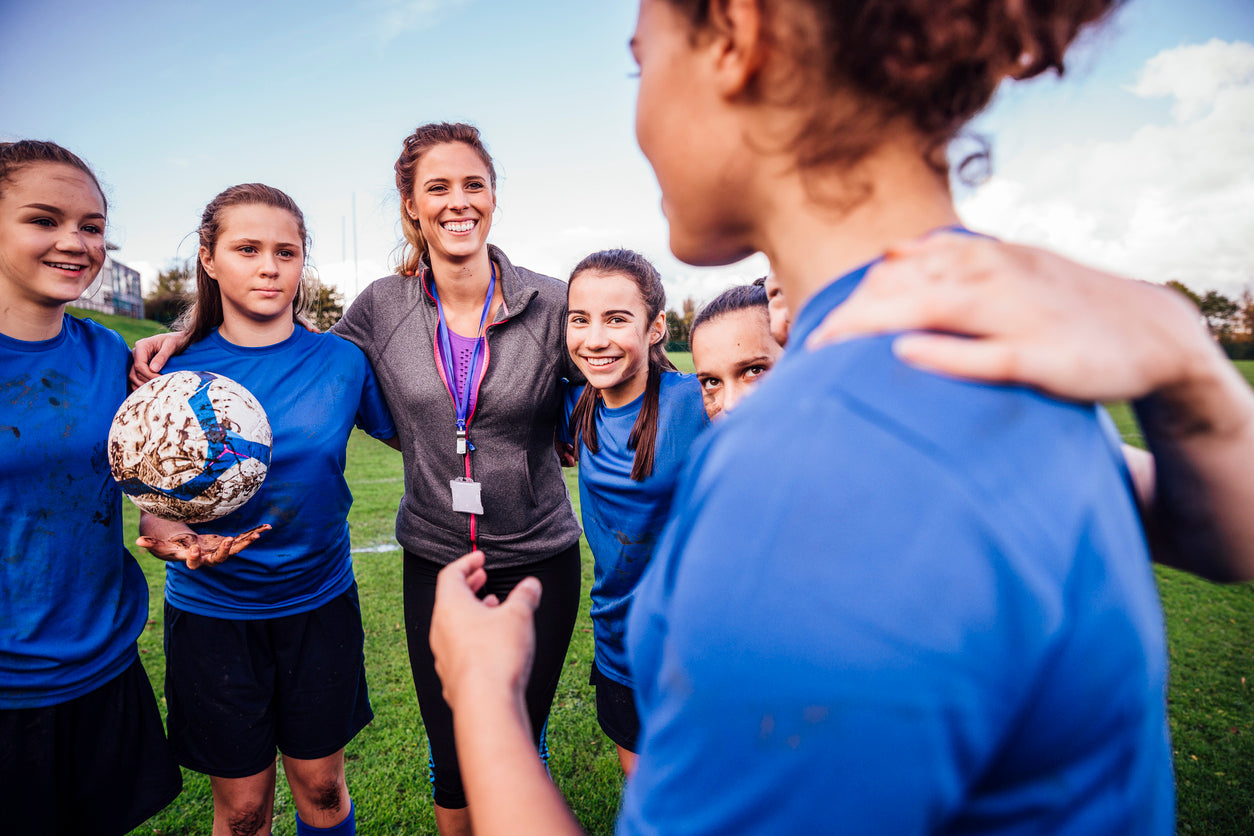 Teenage girls warming up during rugby training with their coach. Their arms are around each other as they stand talking.