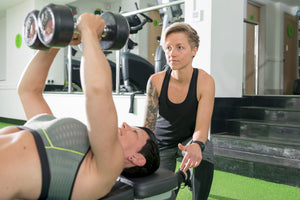 A personal trainer assisting a gym goer lifting weights while lying down.