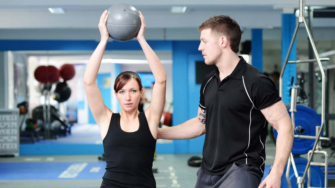 A personal trainer helping a client with their form while lifting a weighted ball